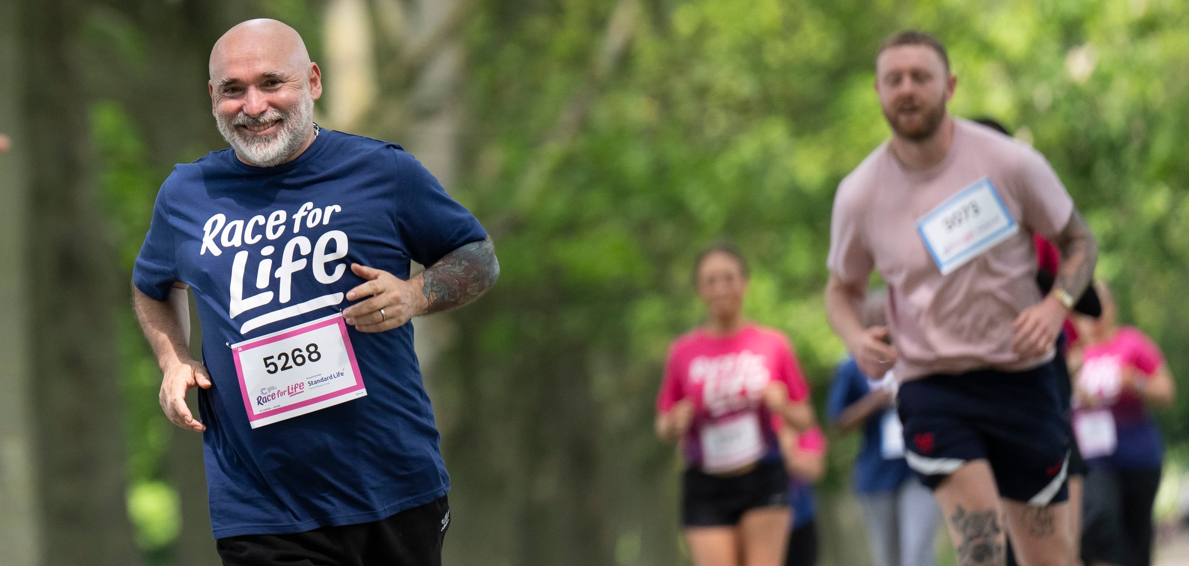 A man is running in park and wearing a blue T-shirt, while taking part in a Race for Life 10K event. He looks happy.
