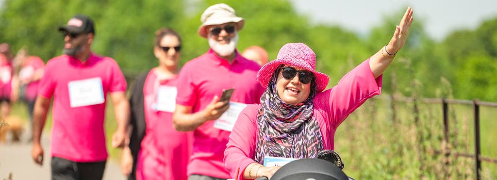 A woman wearing a pink hat and sunglasses raising her hand whilst pushing buggy at 3k race for life event.
