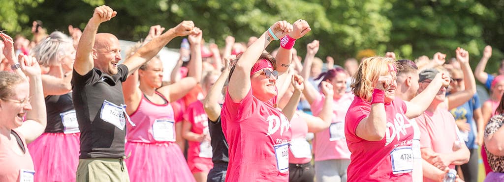 A crowd of Race for Life participants are smiling with their hands in the air. They're warming up before the race begins.